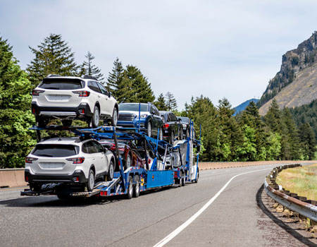 White compact car hauler semi-truck transporting vehicles on a two-level hydraulic trailer along a curving highway through a green mountain forest, used for open auto transport services